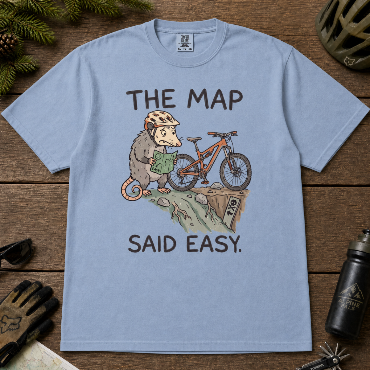 Flat lay of a washed denim blue t-shirt with a humorous mountain biking design showing a nervous opossum, an orange bike, and a dangerously steep trail.