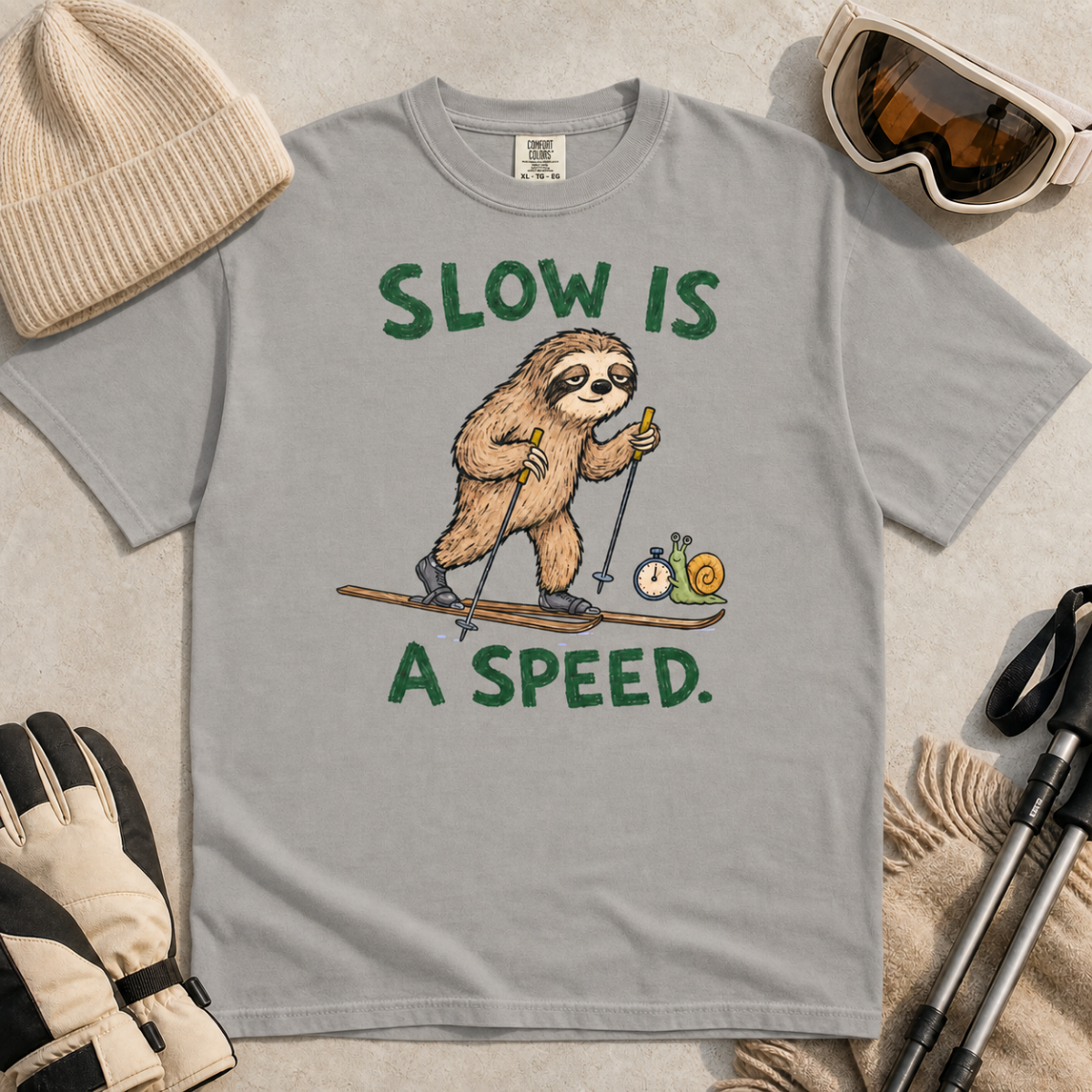 Flat lay of a grey t-shirt with a humorous sloth skiing graphic, a stopwatch-holding snail, and bold green “Slow Is a Speed.” text.