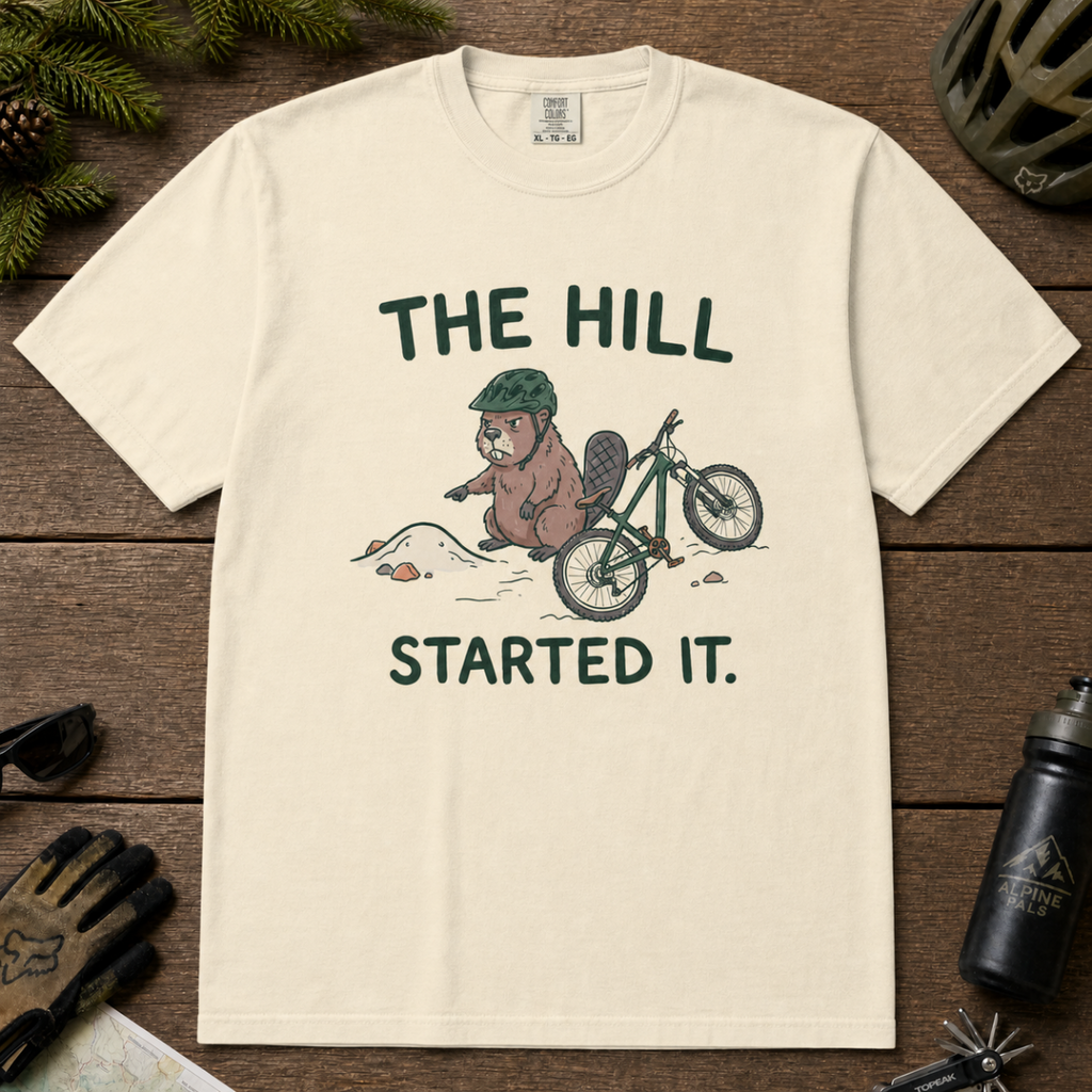Flat lay of an ivory white t-shirt with a humorous mountain biking design showing an annoyed beaver blaming a dirt hill beside its bike.