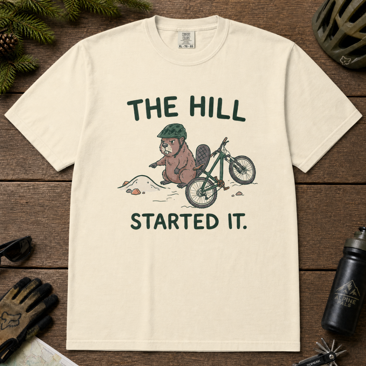 Flat lay of an ivory white t-shirt with a humorous mountain biking design showing an annoyed beaver blaming a dirt hill beside its bike.