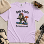 Flat lay of an orchid t-shirt featuring a frustrated badger snowboarder removing one foot from its binding, with “Born to Shred. Forced to Unstrap.” text.