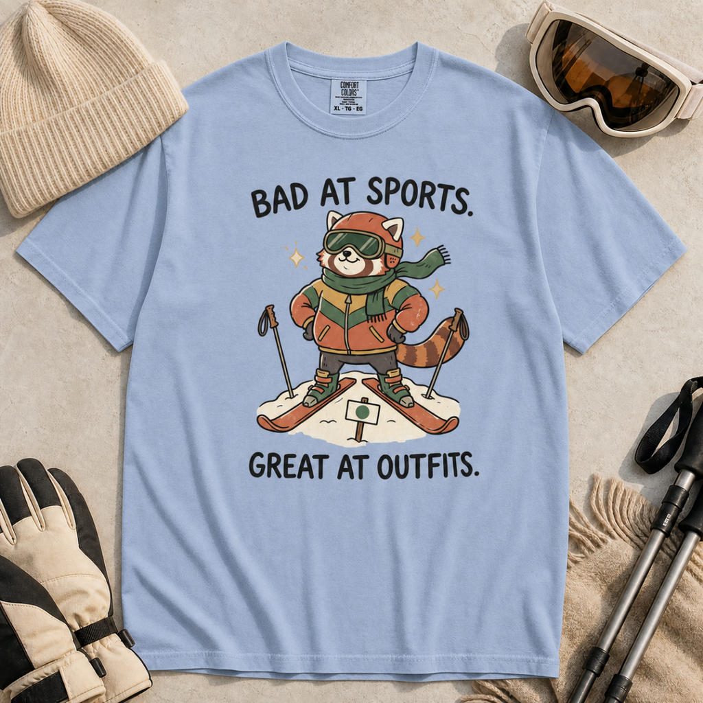 Flat lay of a washed denim blue t-shirt with a humorous red panda ski design celebrating fashionable gear over athletic ability.