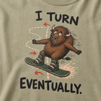 Close-up of a khaki t-shirt showing a wide-eyed bison struggling to steer a snowboard, with directional arrows and “I Turn Eventually.” text.