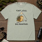 Flat lay of a bay green t-shirt with a playful mountain hiking design showing a small backpacking hamster approaching a tall peak.