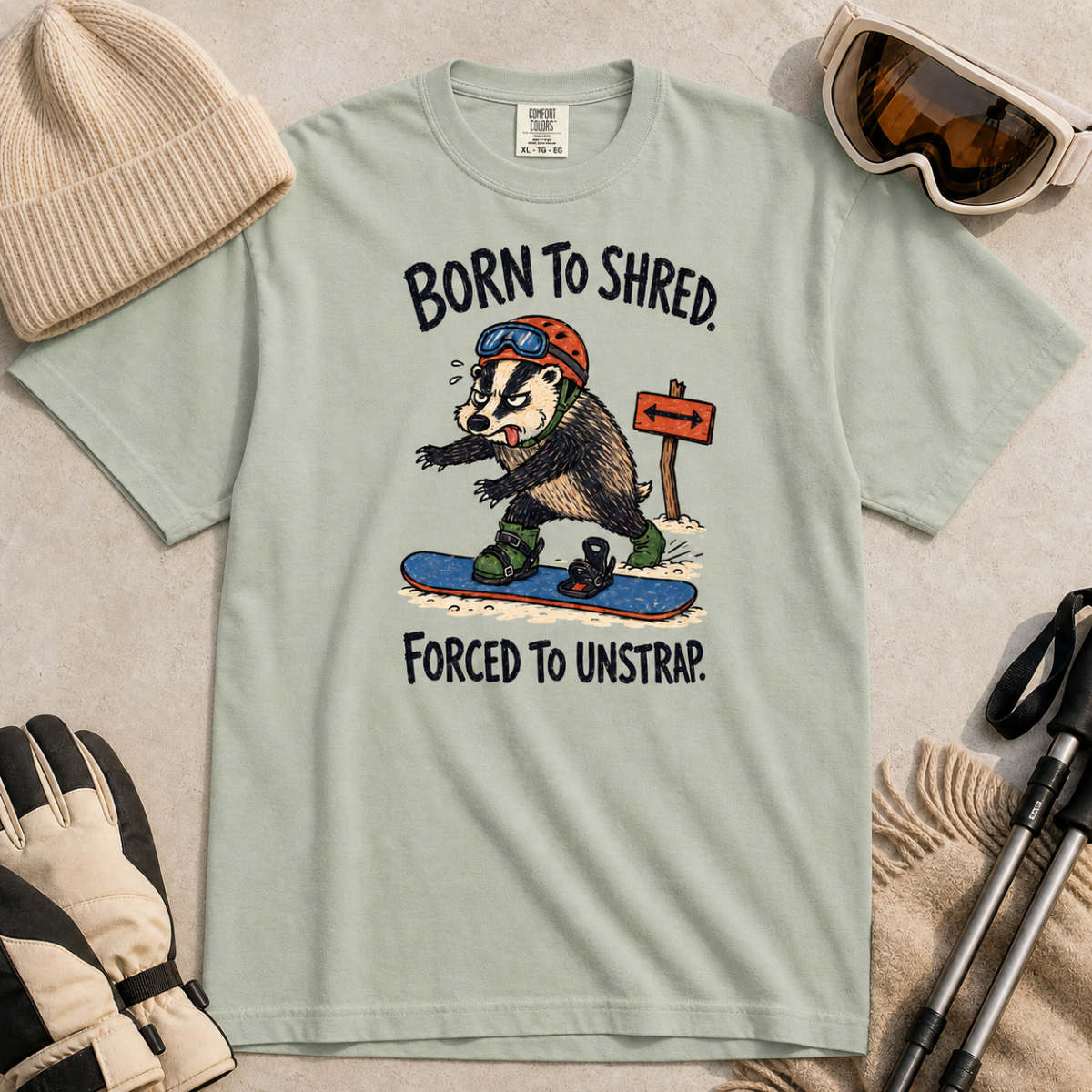 Flat lay of a bay green t-shirt featuring a frustrated badger snowboarder removing one foot from its binding, with “Born to Shred. Forced to Unstrap.” text.