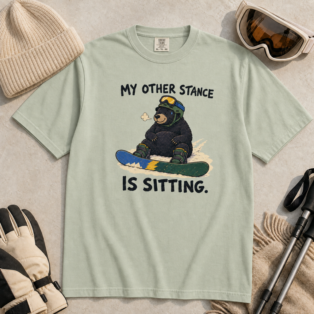 Flat lay of a bay green t-shirt with a humorous snowboarding design showing a resting bear seated behind its board.