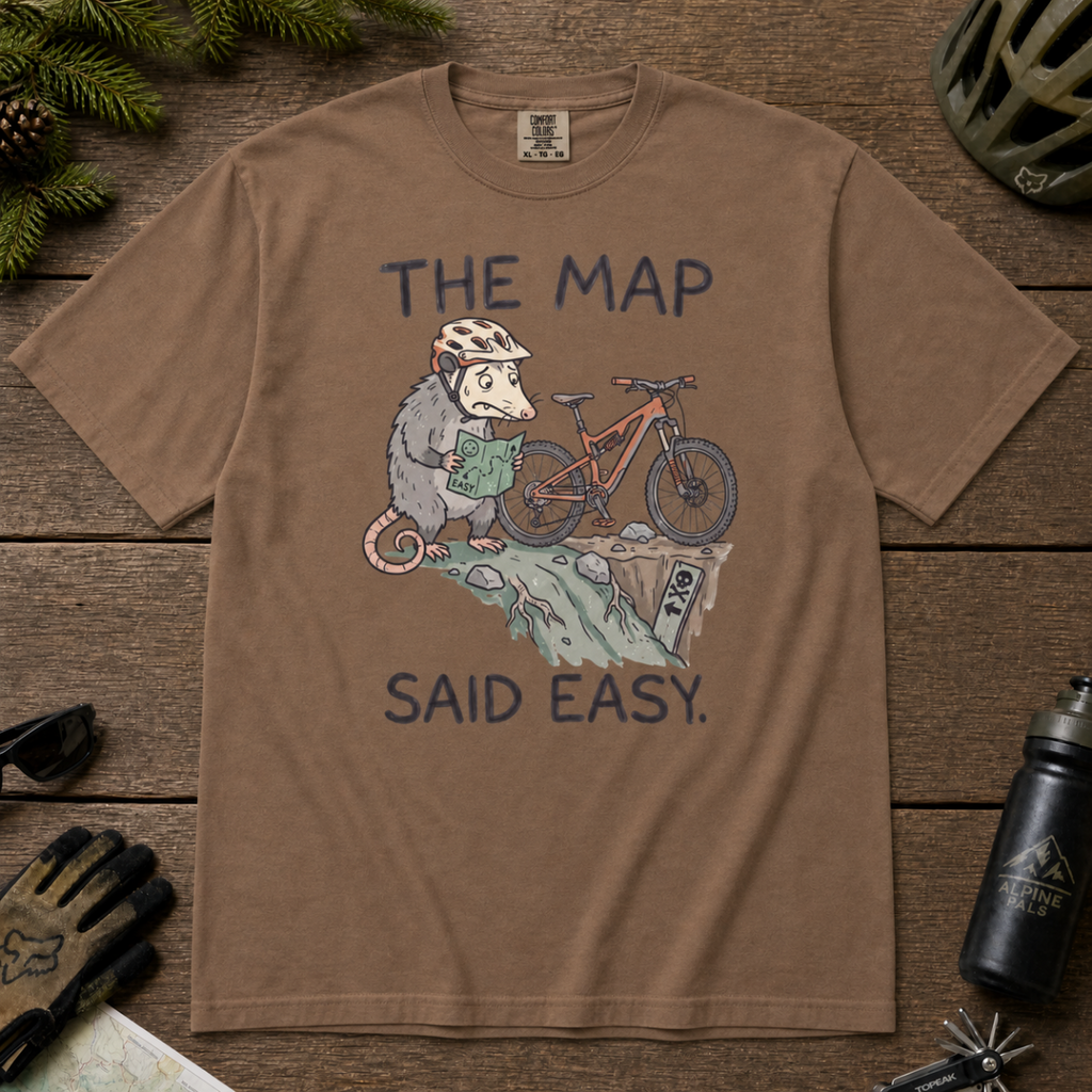 Flat lay of an espresso brown t-shirt with a humorous mountain biking design showing a nervous opossum, an orange bike, and a dangerously steep trail.