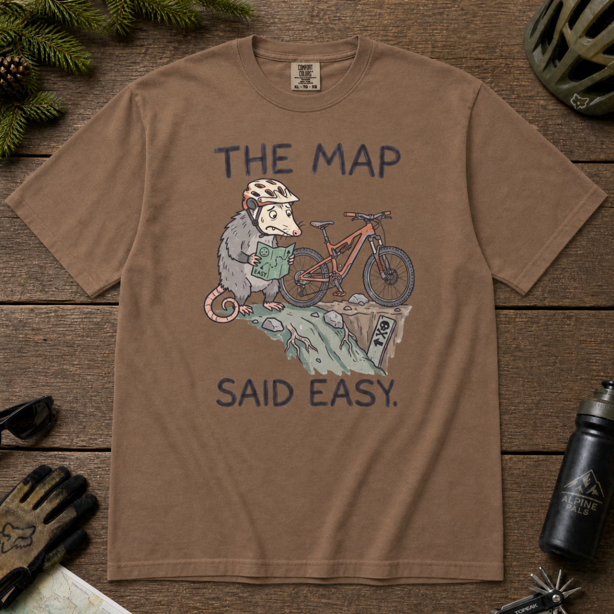 Flat lay of an espresso brown t-shirt with a humorous mountain biking design showing a nervous opossum, an orange bike, and a dangerously steep trail.