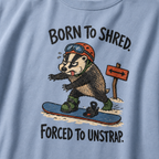 Close-up of a washed denim blue t-shirt showing a tired badger unstrapping from a snowboard near a direction sign, with “Born to Shred. Forced to Unstrap.” text.