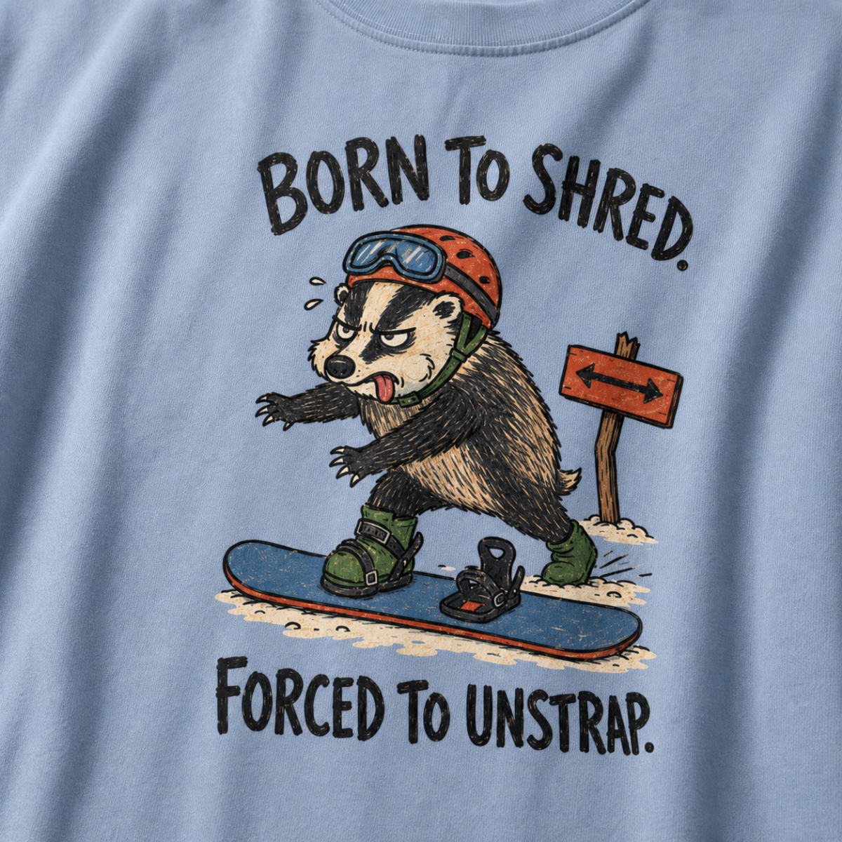 Close-up of a washed denim blue t-shirt showing a tired badger unstrapping from a snowboard near a direction sign, with “Born to Shred. Forced to Unstrap.” text.