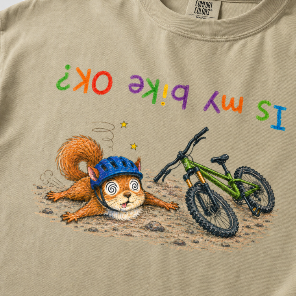 Close-up of a khaki colored t-shirt showing a dazed squirrel after a mountain bike crash beside a green bike, with colourful upside-down “Is My Bike OK?” text.
