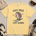 Flat lay of a butter yellow t-shirt featuring a frightened hedgehog skiing at high speed with “Very Fast. Very Scared.” text, styled with winter accessories.