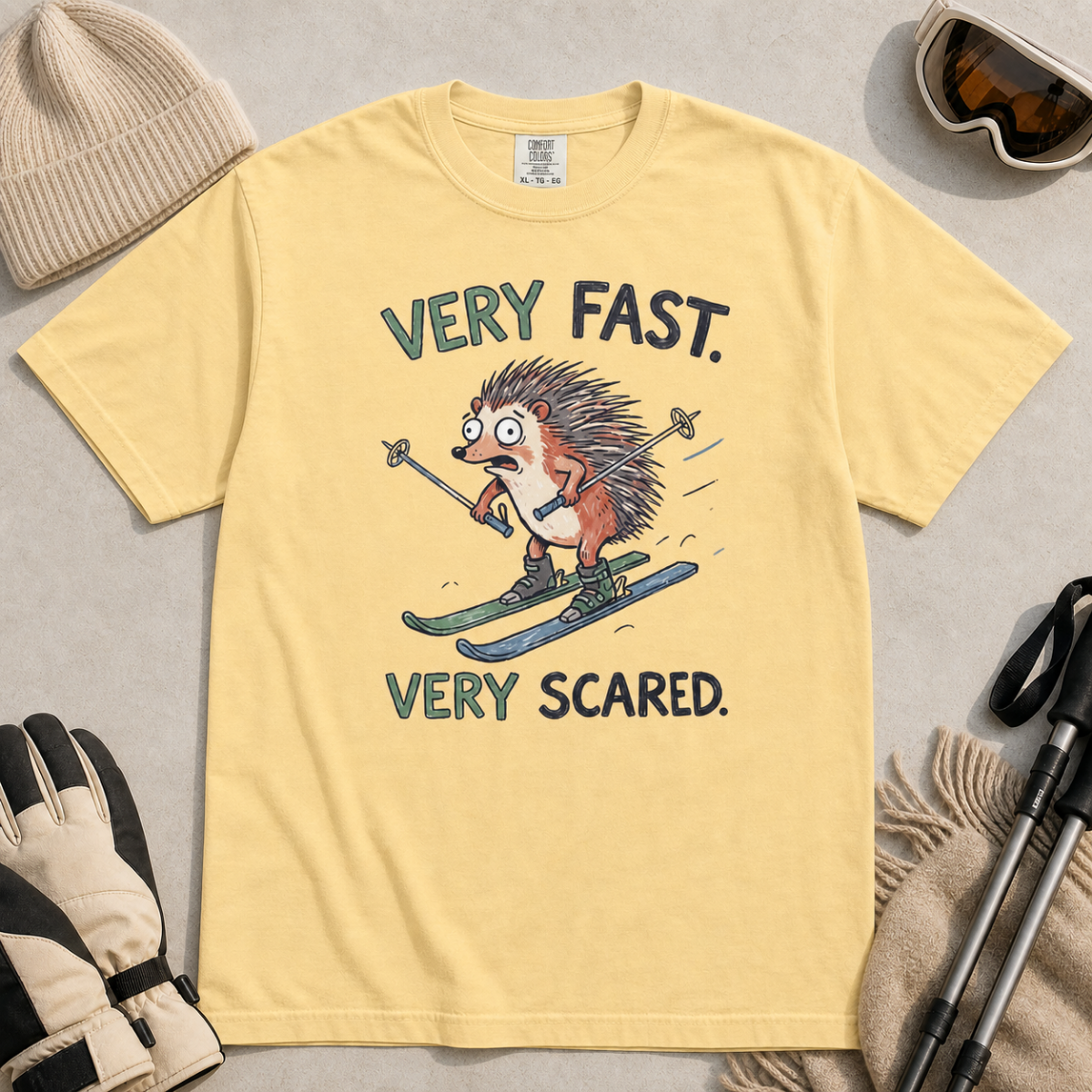 Flat lay of a butter yellow t-shirt featuring a frightened hedgehog skiing at high speed with “Very Fast. Very Scared.” text, styled with winter accessories.