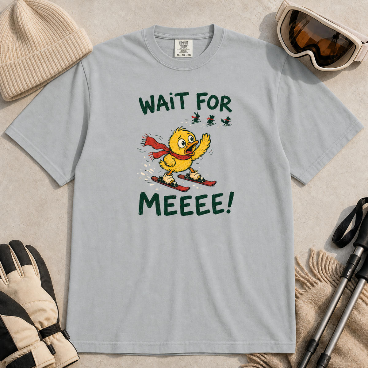 Flat lay of a granite gray t-shirt featuring a panicked duck skiing behind the group with a red scarf and “Wait for Meeee!” text.