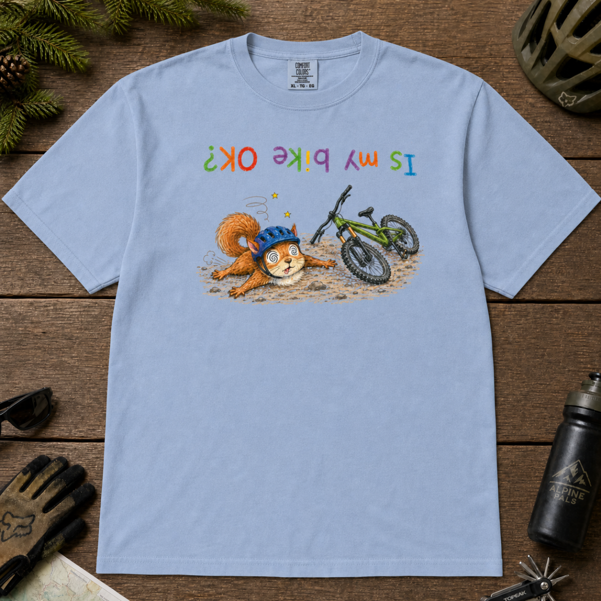 Flat lay of a washed denim blue t-shirt featuring a humorous squirrel mountain bike crash design with an upside-down “Is My Bike OK?” message.