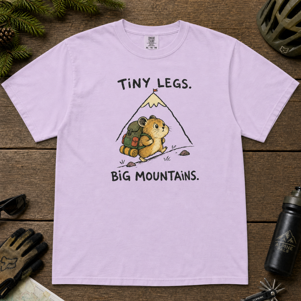 Flat lay of an orchid t-shirt with a playful mountain hiking design showing a small backpacking hamster approaching a tall peak.