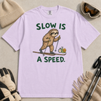 Flat lay of an orchid t-shirt with a humorous sloth skiing graphic, a stopwatch-holding snail, and bold green “Slow Is a Speed.” text.