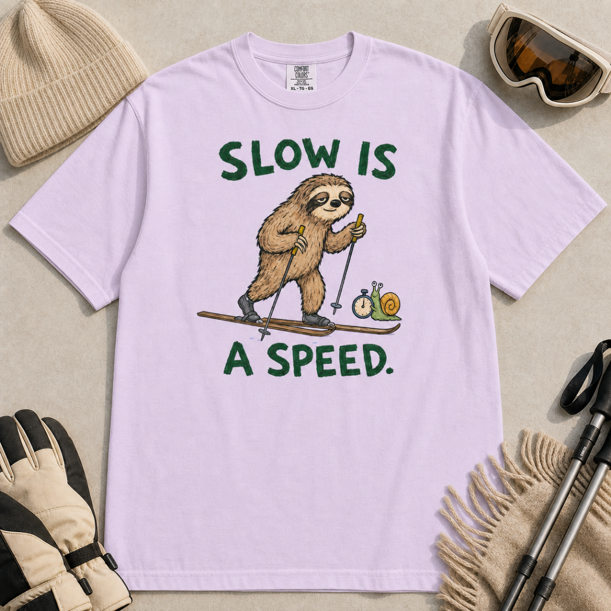 Flat lay of an orchid t-shirt with a humorous sloth skiing graphic, a stopwatch-holding snail, and bold green “Slow Is a Speed.” text.