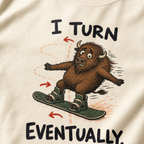 Close-up of an ivory white t-shirt showing a wide-eyed bison struggling to steer a snowboard, with directional arrows and “I Turn Eventually.” text.