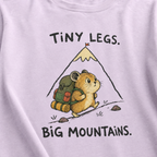 Close-up of an orchid t-shirt featuring a determined hamster hiking uphill with a large backpack beneath “Tiny Legs. Big Mountains.” text.