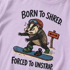 Close-up of an orchid t-shirt showing a tired badger unstrapping from a snowboard near a direction sign, with “Born to Shred. Forced to Unstrap.” text.