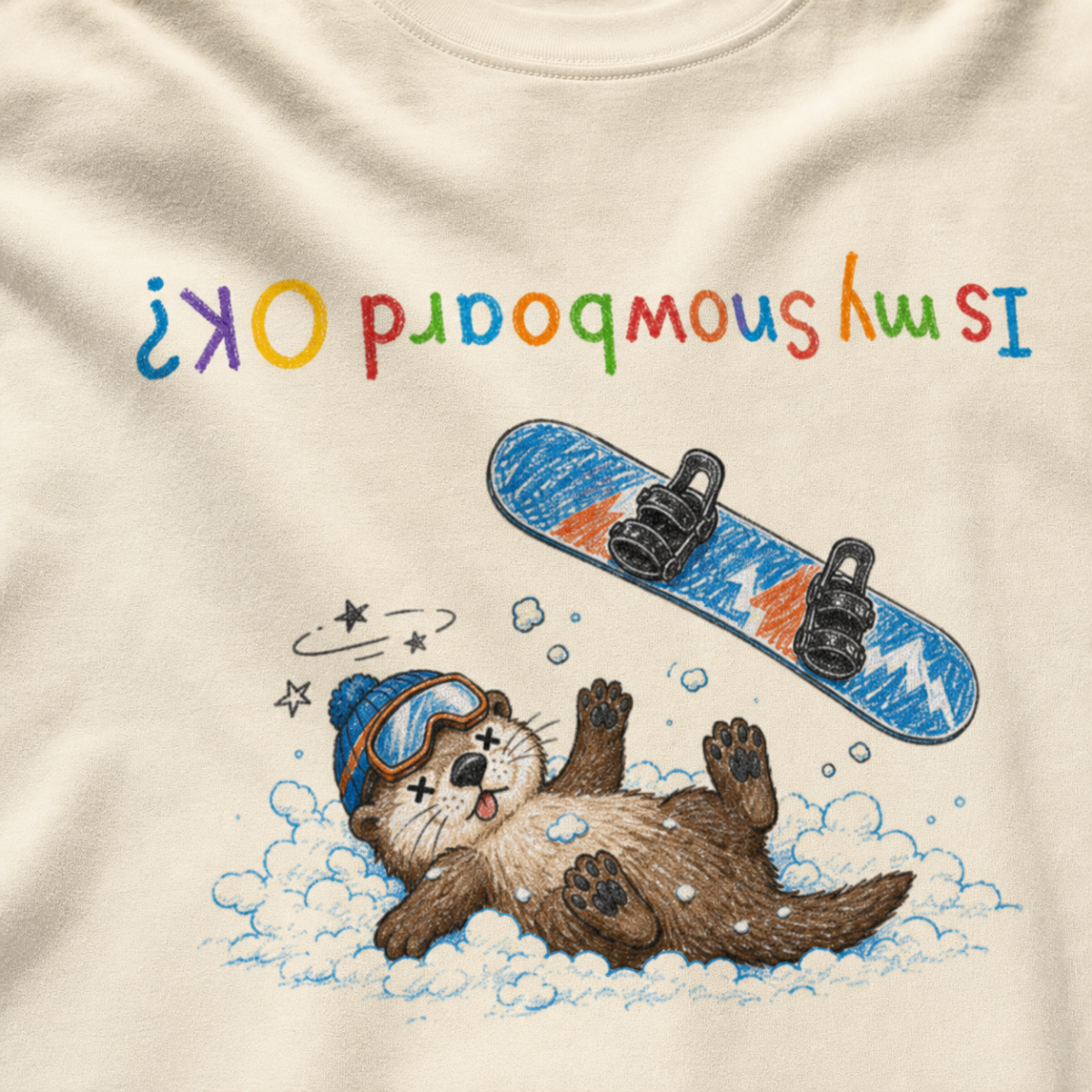 Close-up of an ivory white t-shirt showing a crashed otter in ski goggles with a snowboard flying overhead and colourful upside-down “Is My Snowboard OK?” text.