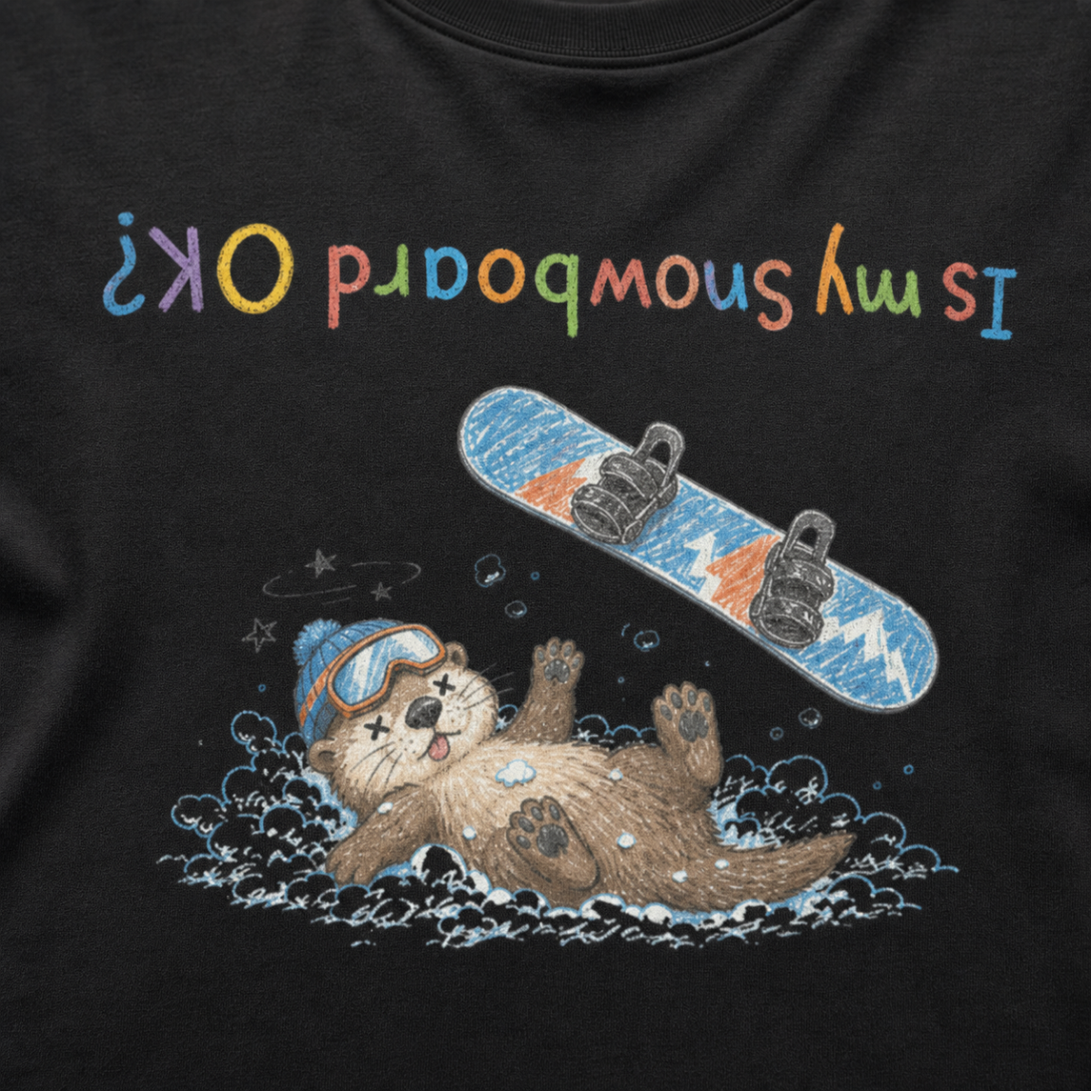 Close-up of a black t-shirt showing a crashed otter in ski goggles with a snowboard flying overhead and colourful upside-down “Is My Snowboard OK?” text.