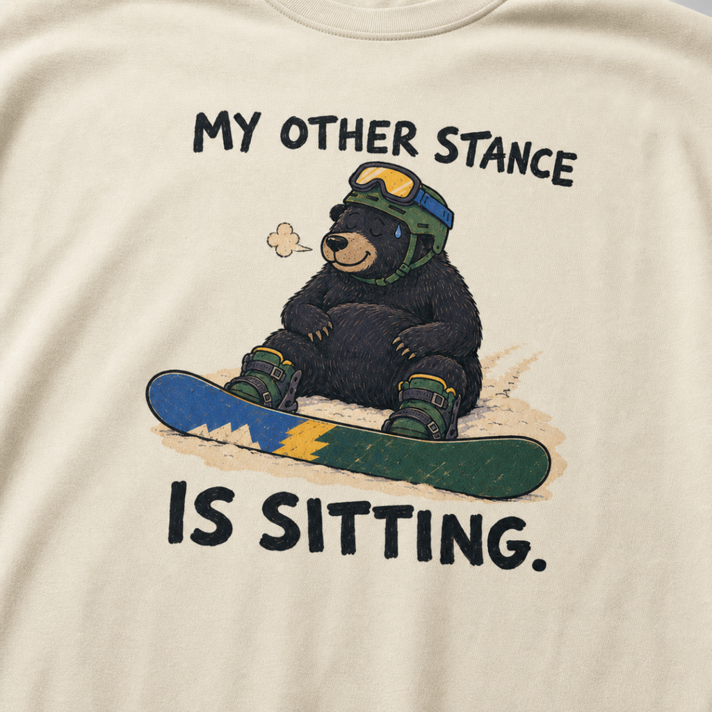 Close-up of an ivory white t-shirt featuring a tired bear sitting in the snow beside a snowboard, with “My Other Stance Is Sitting.” text.