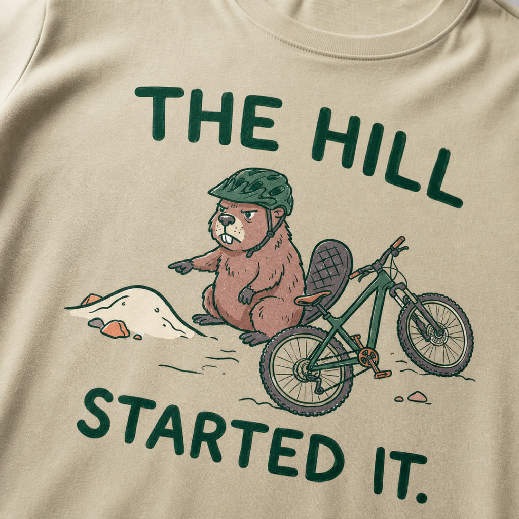 Close-up of a khaki t-shirt featuring a grumpy beaver in a bike helmet pointing at a small dirt hill beside a mountain bike, with “The Hill Started It.” text.