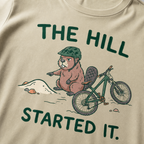 Close-up of a khaki t-shirt featuring a grumpy beaver in a bike helmet pointing at a small dirt hill beside a mountain bike, with “The Hill Started It.” text.
