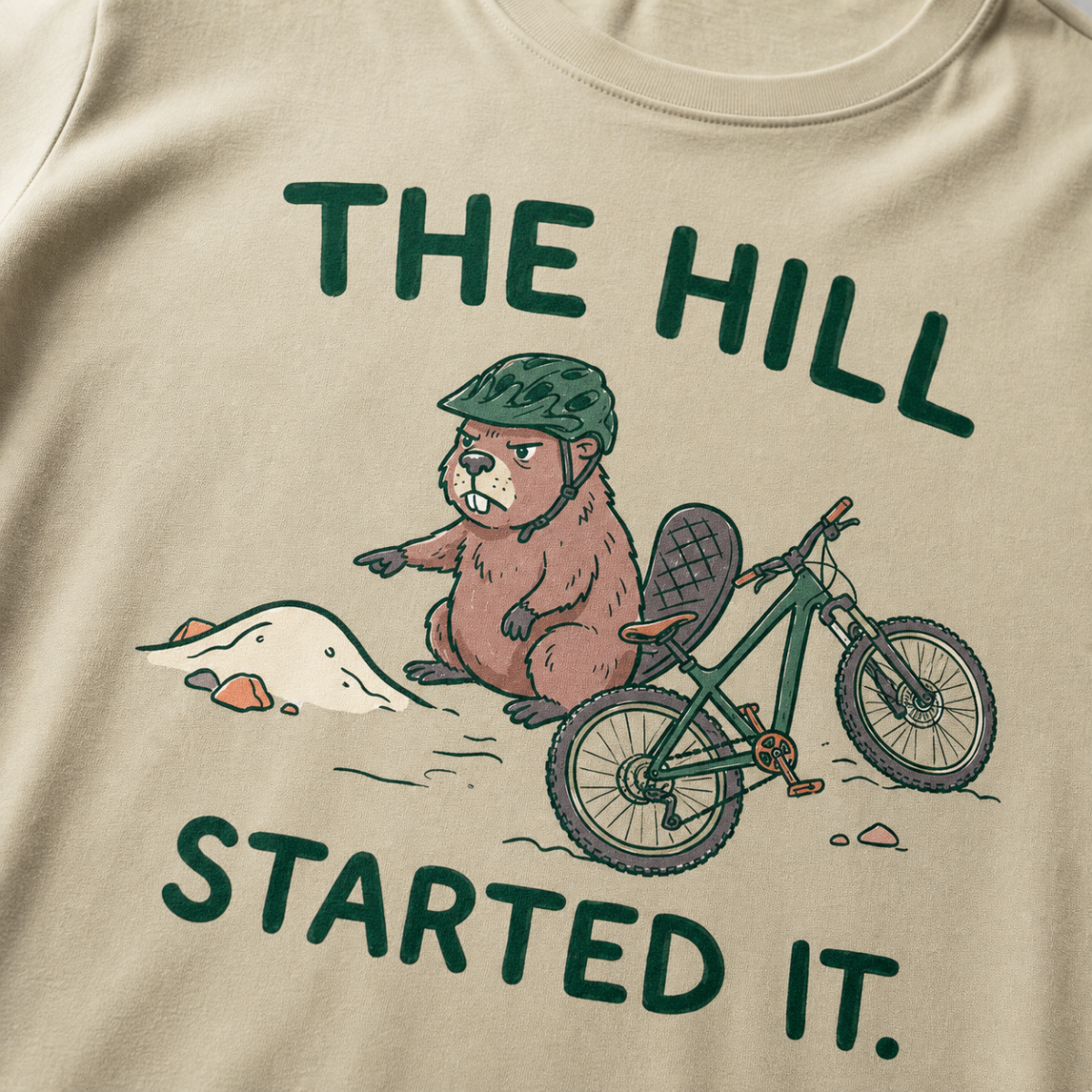 Close-up of a khaki t-shirt featuring a grumpy beaver in a bike helmet pointing at a small dirt hill beside a mountain bike, with “The Hill Started It.” text.