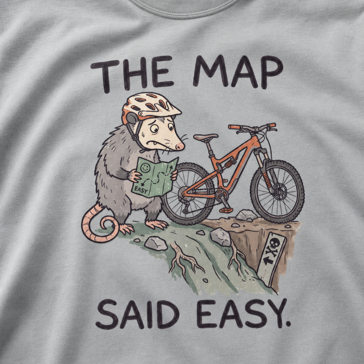 Close-up of a granite gray t-shirt featuring a worried opossum checking an “easy” trail map beside a mountain bike at a steep drop, with “The Map Said Easy.” text.