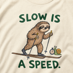 Close-up of an ivory white t-shirt featuring a relaxed sloth cross-country skiing beside a snail with a stopwatch and “Slow Is a Speed.” text.