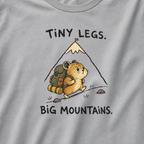 Close-up of a granite gray t-shirt featuring a determined hamster hiking uphill with a large backpack beneath “Tiny Legs. Big Mountains.” text.