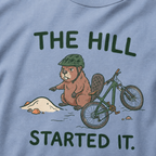 Close-up of a washed denim blue t-shirt featuring a grumpy beaver in a bike helmet pointing at a small dirt hill beside a mountain bike, with “The Hill Started It.” text.