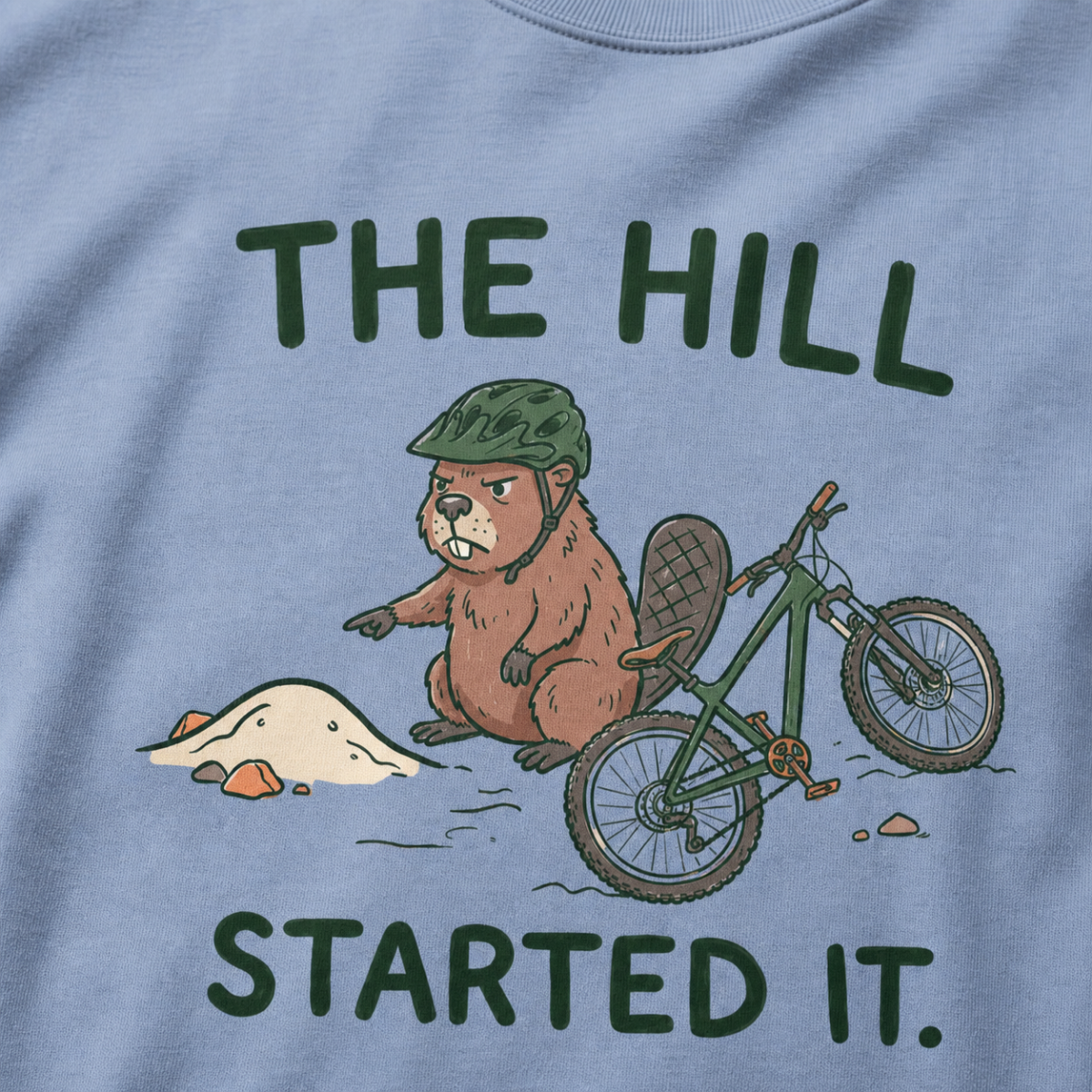 Close-up of a washed denim blue t-shirt featuring a grumpy beaver in a bike helmet pointing at a small dirt hill beside a mountain bike, with “The Hill Started It.” text.