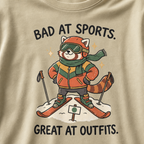 Close-up of a khaki t-shirt featuring a stylish red panda skier posing in coordinated winter gear with “Bad at Sports. Great at Outfits.” text.