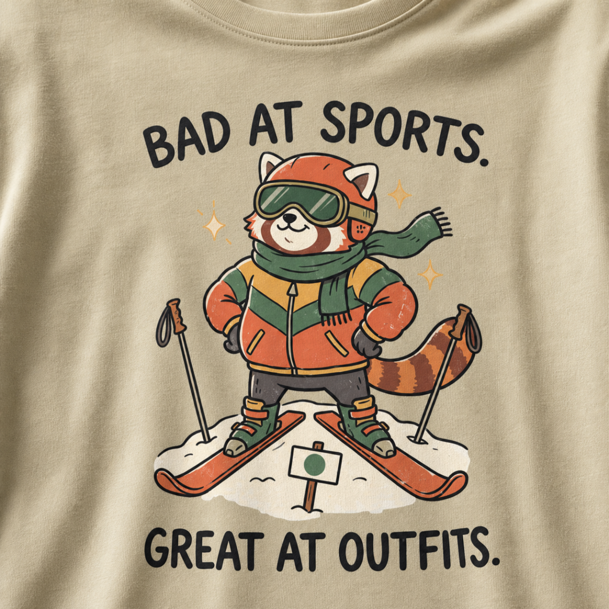 Close-up of a khaki t-shirt featuring a stylish red panda skier posing in coordinated winter gear with “Bad at Sports. Great at Outfits.” text.