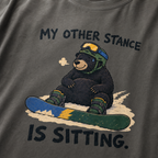 Close-up of a pepper gray t-shirt featuring a tired bear sitting in the snow beside a snowboard, with “My Other Stance Is Sitting.” text.