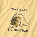 Close-up of a butter yellow t-shirt featuring a determined hamster hiking uphill with a large backpack beneath “Tiny Legs. Big Mountains.” text.