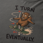 Close-up of a pepper gray t-shirt showing a wide-eyed bison struggling to steer a snowboard, with directional arrows and “I Turn Eventually.” text.