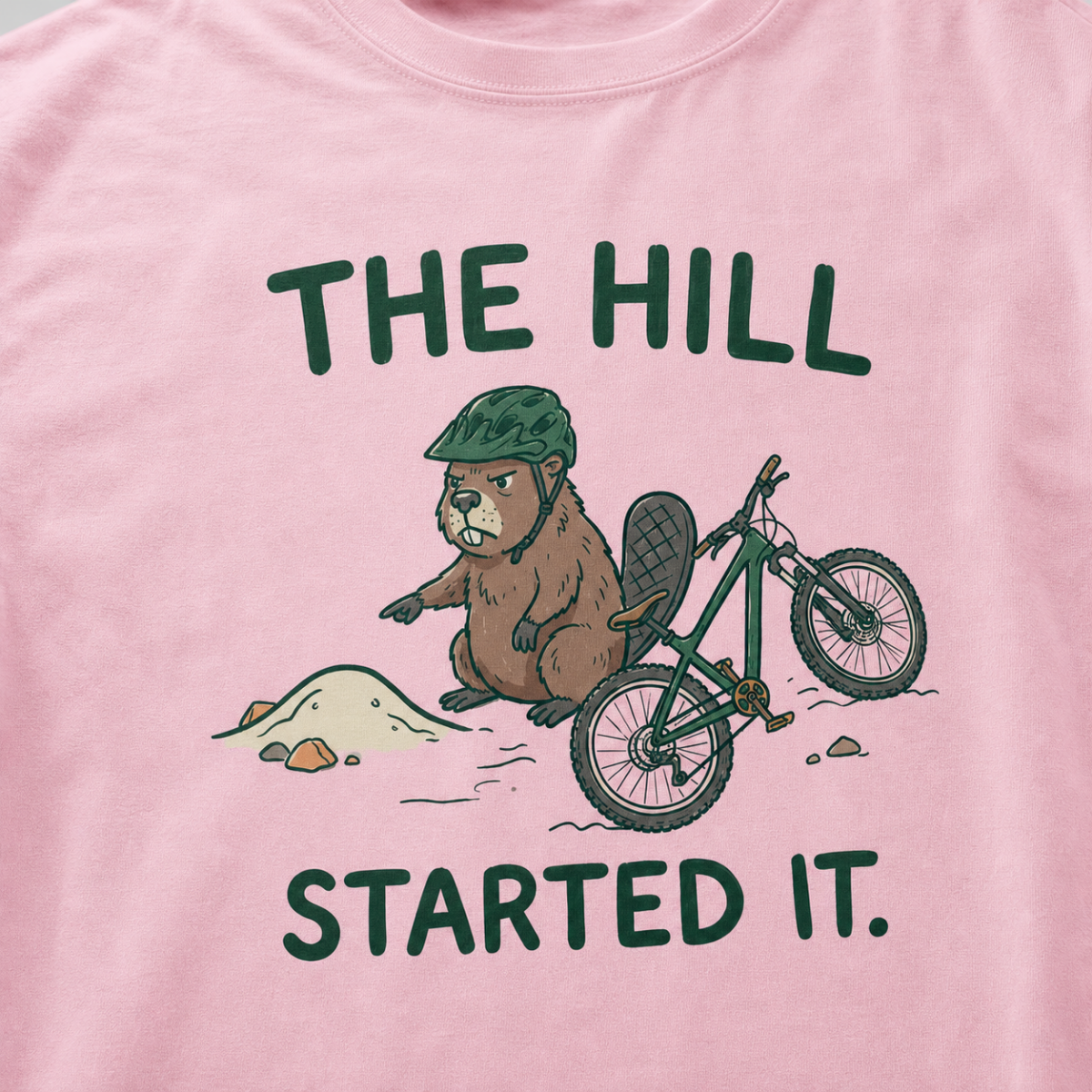Close-up of a blossom pink t-shirt featuring a grumpy beaver in a bike helmet pointing at a small dirt hill beside a mountain bike, with “The Hill Started It.” text.