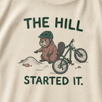 Close-up of an ivory white t-shirt featuring a grumpy beaver in a bike helmet pointing at a small dirt hill beside a mountain bike, with “The Hill Started It.” text.