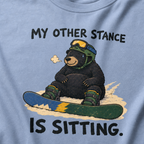 Close-up of a washed denim blue t-shirt featuring a tired bear sitting in the snow beside a snowboard, with “My Other Stance Is Sitting.” text.