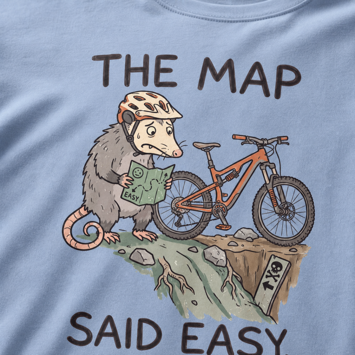 Close-up of a washed denim blue t-shirt featuring a worried opossum checking an “easy” trail map beside a mountain bike at a steep drop, with “The Map Said Easy.” text.
