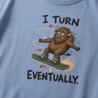 Close-up of a washed denim blue t-shirt showing a wide-eyed bison struggling to steer a snowboard, with directional arrows and “I Turn Eventually.” text.