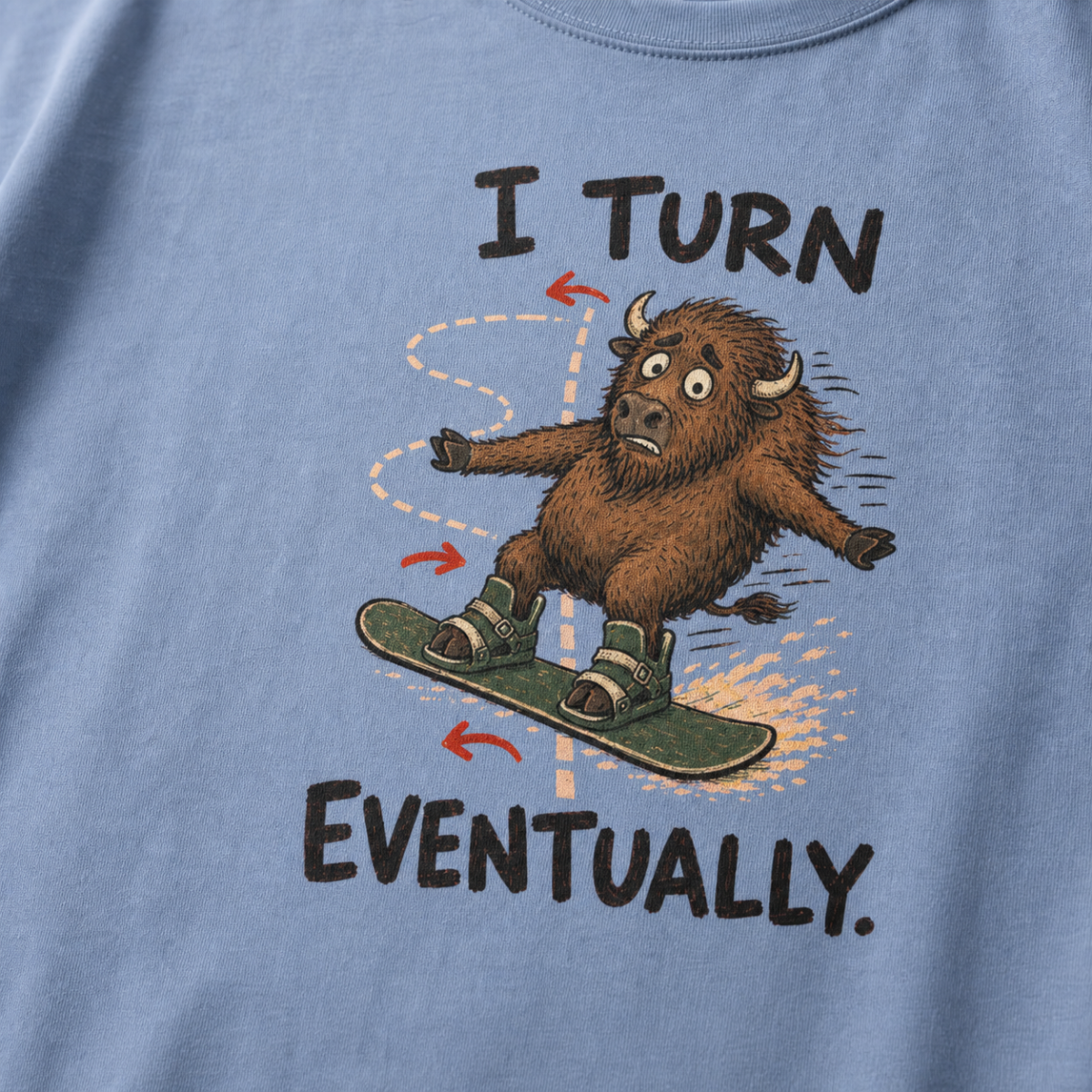 Close-up of a washed denim blue t-shirt showing a wide-eyed bison struggling to steer a snowboard, with directional arrows and “I Turn Eventually.” text.