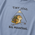 Close-up of a washed denim blue t-shirt featuring a determined hamster hiking uphill with a large backpack beneath “Tiny Legs. Big Mountains.” text.