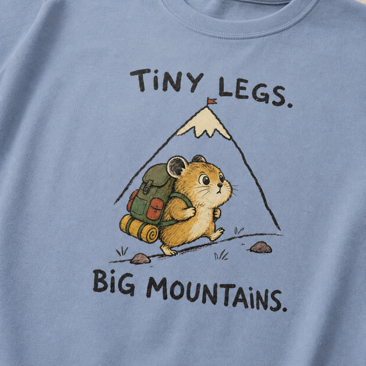 Close-up of a washed denim blue t-shirt featuring a determined hamster hiking uphill with a large backpack beneath “Tiny Legs. Big Mountains.” text.