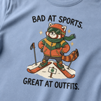 Close-up of a washed denim blue t-shirt featuring a stylish red panda skier posing in coordinated winter gear with “Bad at Sports. Great at Outfits.” text.